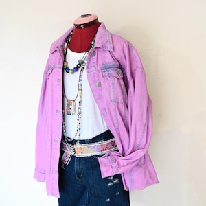 May include: A light purple denim jacket with a white tank top, denim shorts, and a decorative belt. The outfit is accessorized with multiple necklaces. The jacket has button closures and pockets. The shorts have distressed details.
