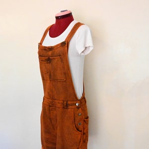 Puede incluir: Un mono de mezclilla marrón con una camiseta blanca debajo. El mono tiene un solo bolsillo delantero y un cierre de botón.