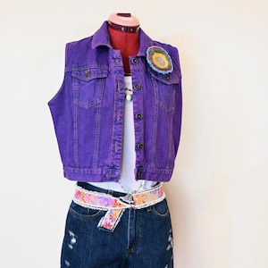 以下が含まれることがあります： 紫色のデニムベストにカラフルな花のパッチ、白いタンクトップ、デニムショーツ、フローラルベルトを組み合わせたファッションアイテム。マネキンに展示されています。 ショーツにはダメージ加工が施されています。