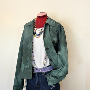 May include: A green jacket with a button-up front, paired with a white t-shirt and denim shorts. The outfit is accessorized with a beaded necklace and a purple belt. The shorts have distressed details.