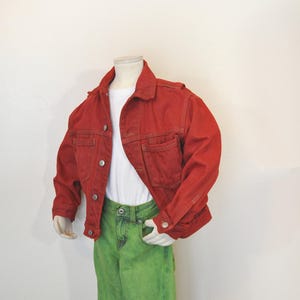 May include: A red denim jacket with silver buttons, worn over a white t-shirt, paired with bright green jeans. The jacket has two chest pockets and a classic collar. The outfit is displayed on a mannequin.