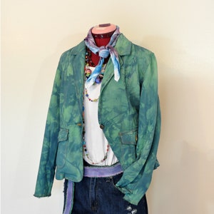 Op de afbeelding: Een groen en blauw tie-dye blazer met een witte top, denim shorts en kleurrijke kralenkettingen. Een blauwe en paarse sjaal is om de nek van de mannequin geknoopt. De shorts hebben distressed details en een paarse riem.