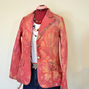 Op de afbeelding: Een rode blazer met een uniek patroon, gecombineerd met een witte top en een spijkerbroekshort, wordt getoond op een mannequin. De blazer heeft twee zakken en een sluiting met twee knopen. Een kleurrijke kralenketting en riem maken de outfit af. De mannequin staat op een rode standaard.