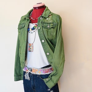 May include: An olive green distressed denim jacket with button closures and chest pockets. The jacket is paired with a white ribbed tank top, denim shorts, and a floral patterned belt. A mannequin head displays the outfit with layered necklaces.