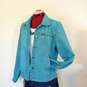 May include: A teal blue denim jacket with a button closure and a small logo on the left chest. The jacket is worn over a white shirt and blue denim shorts.