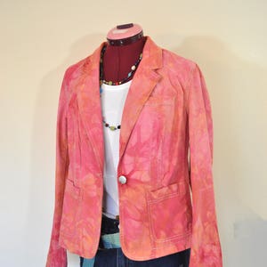Op de afbeelding: Een roze tie-dye blazer met een witte knoop en twee zakken. De blazer is op een mannequin en wordt gecombineerd met een wit T-shirt, een blauwe riem en een spijkerbroek.