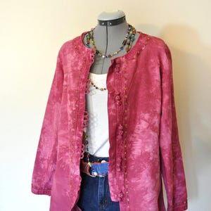 May include: A pink tie-dye jacket with gold-colored embellishments and buttons. The jacket is paired with a white top, denim shorts, and a beaded necklace. A blue belt and a blue fabric detail are also visible.