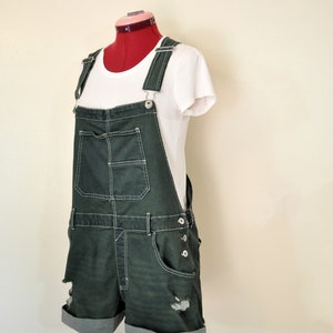 May include: Dark green denim overall shorts with white stitching and distressed details. The shorts have rolled cuffs and are paired with a white t-shirt. The outfit is displayed on a red dress form.