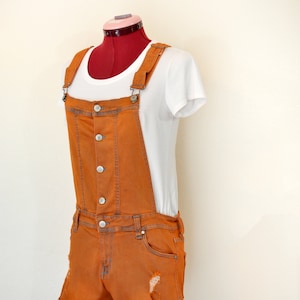 Puede incluir: Pantalones cortos de peto vaqueros naranjas con detalles desgastados, combinados con una camiseta blanca. Los pantalones cortos tienen una botonadura frontal y dobladillos. El conjunto se muestra en un maniquí rojo.