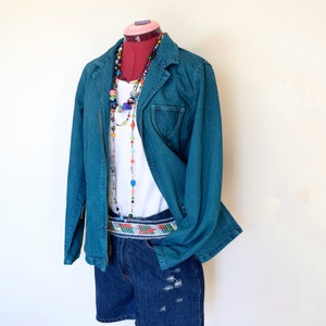 May include: A teal denim jacket with a zipper closure and a chest pocket is displayed on a mannequin. The jacket is paired with a white top, denim shorts, and colorful beaded necklaces. A patterned belt adds a pop of color.