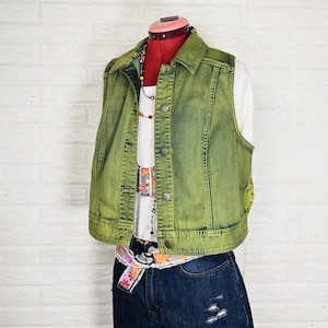 May include: A sleeveless olive green denim vest with a collared neckline and metal button closures, worn over a white ribbed top and dark blue denim shorts with distressed details. A colorful beaded necklace and a patterned fabric belt are visible.