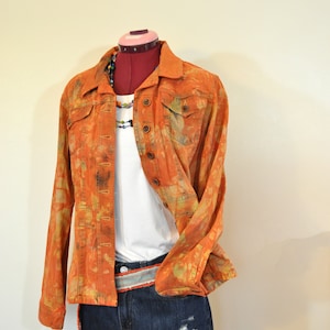 Puede incluir: Una chaqueta vaquera con un estampado tie-dye en tonos naranja y amarillo. La chaqueta se lleva sobre una camiseta blanca y unos pantalones cortos de mezclilla azul con un cinturón marrón.