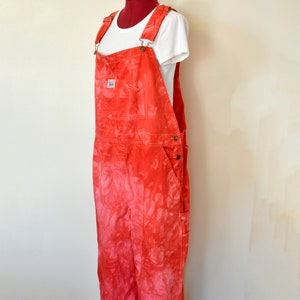 Könnte beinhalten: Rote Tie-Dye-Latzhose mit verstellbaren Trägern und mehreren Taschen. Die Latzhose wird auf einer Schaufensterpuppe mit einem weißen T-Shirt präsentiert. Die Latzhose hat ein verwaschenes, strukturiertes Aussehen. Ein kleines Etikett mit dem Wort "BLUE" ist auf der Vordertasche sichtbar.