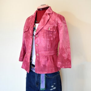 May include: A pink blazer with three-quarter sleeves, button closures, and two front pockets. The blazer is paired with a white top, denim shorts, and a colorful belt. A necklace is also visible.
