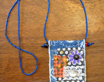 May include: A handmade denim pendant necklace with a blue cord. The pendant has a patchwork design with a purple flower, white lace, and a button. The denim is decorated with white polka dots and a floral pattern.