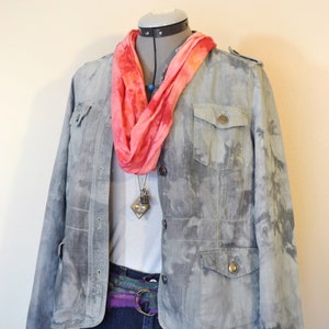 May include: A gray jacket with a tie-dye effect, featuring two chest pockets and button closures. A pink and orange scarf is draped around the neck. The outfit includes a white shirt, dark blue jeans, and a decorative belt.