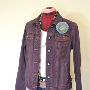 May include: A plum-colored denim jacket with button closures, chest pockets, and a collar. The jacket is worn over a white shirt and blue jeans. A colorful brooch and beaded necklaces add accents to the outfit.