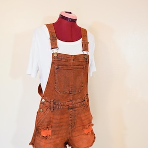 Puede incluir: Pantalones cortos de peto vaqueros naranjas con detalles desgastados. Los pantalones cortos tienen un bolsillo delantero, tirantes ajustables y un dobladillo deshilachado. Se ve una camiseta blanca debajo.