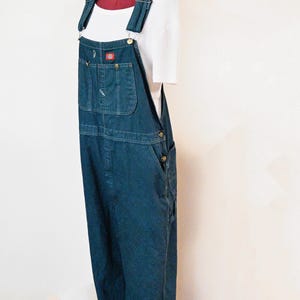 Può includere: Salopette in denim blu scuro con spalline regolabili e multiple tasche. La salopette presenta un design classico con una toppa con il logo Dickies sulla tasca sul petto. Sotto è visibile una maglietta bianca.