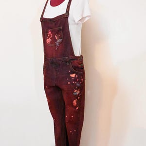 Puede incluir: Un mono de mezclilla rojo oscuro con salpicaduras de pintura en tonos de rosa, naranja y gris. El mono tiene un diseño clásico con tirantes ajustables y un bolsillo delantero.