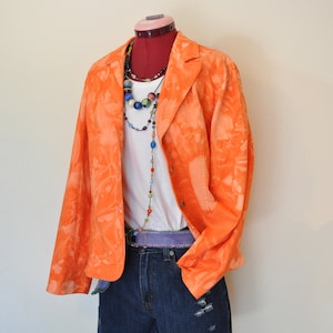 Puede incluir: Un blazer naranja teñido anudado se exhibe en un maniquí, combinado con una camiseta blanca, pantalones cortos de mezclilla y un collar de cuentas. El atuendo incluye un cinturón morado. El maniquí está sobre un soporte rojo.