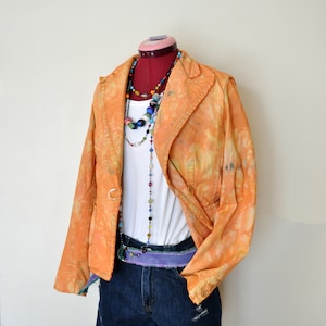 Puede incluir: Una blazer naranja teñida con un top blanco y pantalones cortos de mezclilla. El atuendo se complementa con collares de cuentas y un cinturón morado. El maniquí está sobre un soporte rojo.