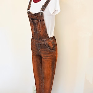 Puede incluir: Pantalón vaquero naranja con tirantes, camiseta blanca y bandana de colores. El pantalón tiene un bolsillo delantero y tirantes ajustables.