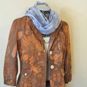 Puede incluir: Una blazer marrón con patrones anaranjados en forma de hojas, botones blancos y una bufanda azul claro. La blazer se combina con una camiseta blanca y una falda vaquera. También se ve un collar colorido.