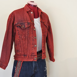 May include: A red denim jacket with a white t-shirt underneath and a dark blue denim shorts with a multi-colored belt.