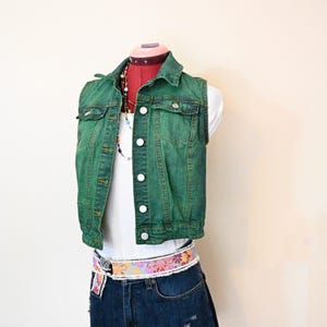 Op de afbeelding: Een groene spijkerweste met witte knopen wordt gedragen over een witte tanktop. De outfit omvat een distressed spijkerbroek en een bloemenriem. Een kleurrijke kralenketting voegt een vleugje kleur toe aan de ensemble.