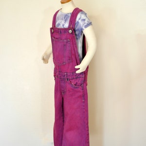 May include: A mannequin displays a pair of bright magenta denim overalls with a front pocket and adjustable straps. The overalls are paired with a tie-dye t-shirt. The outfit is casual and stylish.