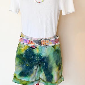 Puede incluir: Pantalones cortos de mezclilla teñidos con tie-dye verde y azul con un cinturón de tela floral multicolor.