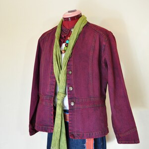 Puede incluir: Chaqueta vaquera burdeos con botones plateados, combinada con una bufanda verde y una falda vaquera, exhibida en un maniquí. El conjunto sugiere un estilo casual y en capas. Un collar de cuentas coloridas añade un toque vibrante.