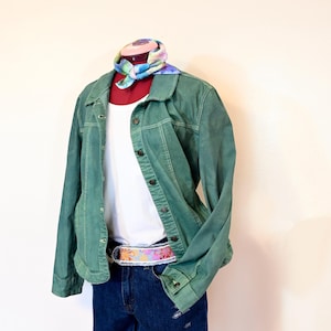 May include: A green denim jacket with a white t-shirt underneath and a floral patterned belt. The jacket has a button-up front and long sleeves. The model is wearing blue denim shorts.
