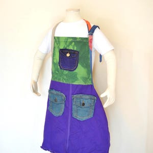Puede incluir: Delantal hecho a mano con un diseño de patchwork único. La sección superior es verde con efecto tie-dye y un bolsillo de mezclilla azul. La sección inferior es morada con dos bolsillos de mezclilla. Con el texto "Apron #154 Size 8-12".
