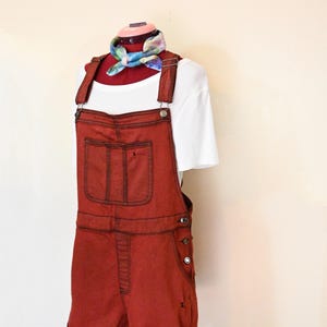 Könnte beinhalten: Rostfarbene Jeans-Latzhose mit dunkelbraunen Nähten und einer Vordertasche. Die Latzhose wird mit einem weißen T-Shirt und einem bunten Schal kombiniert. Das Outfit wird auf einer Schaufensterpuppe präsentiert.