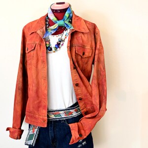 Puede incluir: Una chaqueta vaquera de color óxido con una camiseta blanca, un collar de cuentas de colores y un pañuelo de bandana teñido anudado. La chaqueta tiene un cierre de botones en la parte delantera y dos bolsillos en el pecho. La modelo lleva pantalones cortos de mezclilla azul con un cinturón tejido de colores.