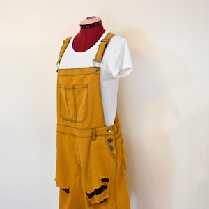 Op de afbeelding: Mosterdgele denim overall met distressed details en een wit T-shirt eronder. De overall heeft verstelbare banden en een enkele voorzak.