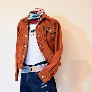 Puede incluir: Una chaqueta vaquera de color óxido con una camiseta blanca debajo. La chaqueta tiene un cinturón con estampado floral y un collar de cuentas multicolores. La modelo lleva pantalones cortos de mezclilla azul.