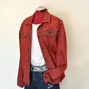 May include: A rust-colored denim jacket with button closures, worn over a white t-shirt and dark blue denim shorts. A colorful patterned belt accents the waist. The outfit is displayed on a mannequin.