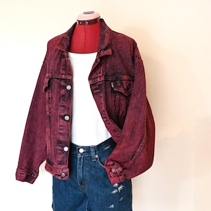 May include: A burgundy denim jacket with a faded, acid-wash effect, featuring silver buttons and two chest pockets. The jacket is paired with a white tank top and blue denim shorts. The outfit is displayed on a red dress form.