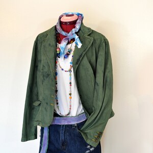 Op de afbeelding: Een outfit op een paspop bestaande uit een groene spijkerjas, een witte top en een spijkerbroekshort. De outfit is geaccentueerd met een kleurrijke sjaal, kralenkettingen en een paarse riem. De shorts hebben distressed details.