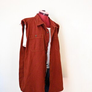 May include: A sleeveless rust-colored denim vest featuring a classic collar, a button-down front, and two chest pockets. The armholes have a raw, frayed edge finish. The vest is layered over a white ribbed tank top and dark blue denim jeans.