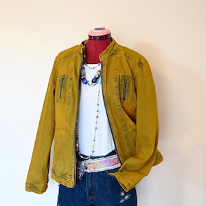 Op de afbeelding: Een mosterdgele denimjas met een wit T-shirt en een blauwe denimshort. De jas heeft een ritssluiting en meerdere zakken. De short heeft een bloemenprint riem.