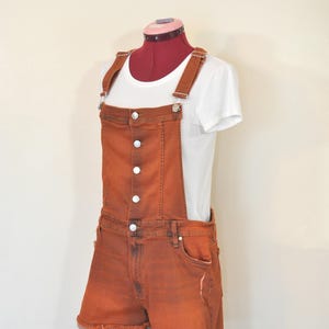 Puede incluir: Pantalones cortos con peto de mezclilla naranja quemado con botones blancos y un bolsillo en el pecho. Los pantalones cortos tienen un dobladillo desgastado y se combinan con una camiseta blanca. El atuendo se muestra en un maniquí.