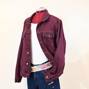 Puede incluir: Chaqueta vaquera burdeos con botones, usada sobre una camiseta sin mangas blanca de canalé y pantalones cortos vaqueros azules. Un cinturón con estampado floral añade un toque de color. El conjunto se exhibe en un maniquí.