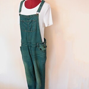 Op de afbeelding: Een teal denim overall met een voorzak en verstelbare bandjes, gecombineerd met een wit geribbeld shirt met korte mouwen. De outfit wordt getoond op een rode paspop.