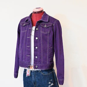 以下が含まれることがあります： ボタンとポケットが付いた紫色のデニムジャケット。白いトップス、デニムショーツ、フローラルベルトと組み合わせています。マネキンに飾られたコーディネートです。