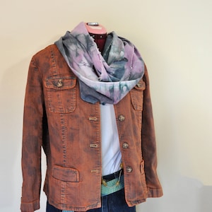 Puede incluir: Una chaqueta vaquera de color óxido con una camisa blanca debajo y una bufanda azul y morada. La chaqueta tiene un cierre de botones en la parte delantera y dos bolsillos en la parte delantera. La chaqueta se lleva sobre unos pantalones cortos de mezclilla azul.