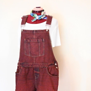 Könnte beinhalten: Burgunderfarbene Jeans-Latzhose-Shorts auf einer Schaufensterpuppe. Die Shorts haben einen ausgefransten Saum und Taschen. Ein weißes T-Shirt und ein farbenfrohes Halstuch werden darunter getragen. Die Latzhosenträger haben silberfarbene Beschläge.